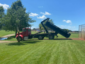 A red roll-off truck from Minnesota Waste and Transfer lifting a black dumpster in a grassy area in Champlin, MN