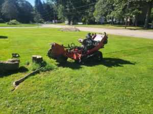 A red stump grinder positioned next to a large tree stump and cut logs on a grassy lawn, ready for service by Grin & Grind Stump Removal LLC in Worcester, MA.
