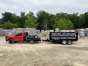 A red truck towing a dump trailer with Mobile Dumpster Solutions branding at a material yard in Chattanooga, TN.