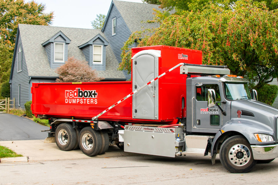 A redbox+ truck delivering a dumpster with a portable toilet to a residential property in Baton Rouge, LA, by redbox+ of Baton Rouge.