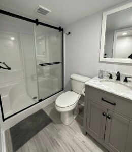 A renovated bathroom with a grey vanity, toilet, and a modern shower-tub combination by Impressive Basements in Westland, MI.