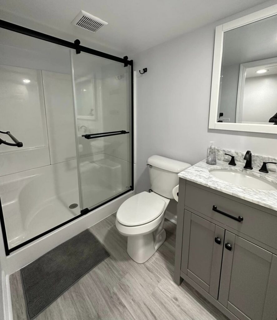 A renovated bathroom with a grey vanity, toilet, and a modern shower-tub combination by Impressive Basements in Westland, MI.