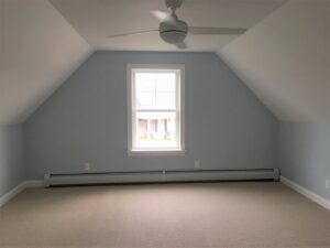 A bright, renovated attic room with light blue walls, a large window, and a ceiling fan by Casco Bay Construction Services, LLC in South Portland, ME.