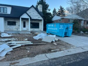 A blue dumpster positioned at a home renovation site with debris, provided by Blue Bin Dumpster Rentals in Salt Lake City, UT.