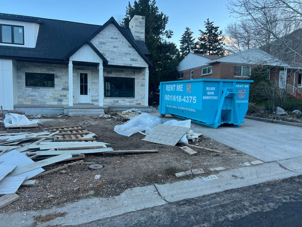 A blue dumpster positioned at a home renovation site with debris, provided by Blue Bin Dumpster Rentals in Salt Lake City, UT.