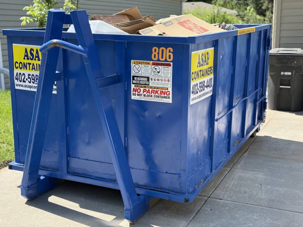 An ASAP Containers inc. blue dumpster filled with cardboard and junk in a residential driveway in Omaha, NE.