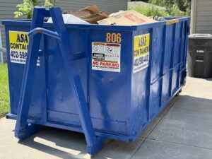 An ASAP Containers inc. blue dumpster filled with cardboard and junk in a residential driveway in Omaha, NE.