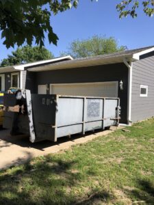 A gray dumpster placed on a residential driveway on a sunny day by NID Dumpster Rental in Nashua, IA.