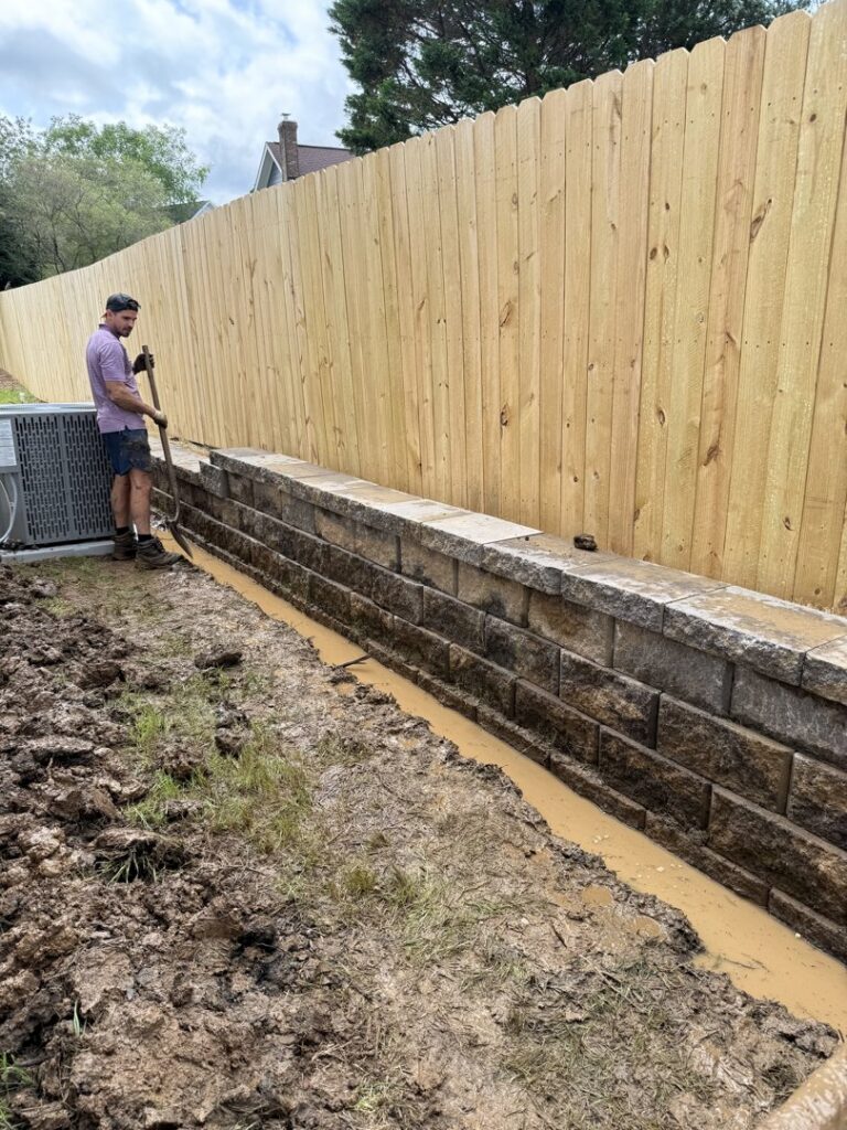 A worker digging a drainage trench next to a newly built retaining wall for Drip Dry Drainage in Cramerton, NC.