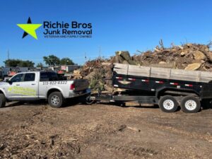 A Richie Bros Junk Removal truck and trailer next to a large pile of tree branches and wood debris in Urbandale, IA.