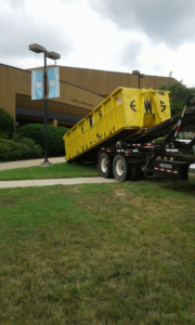 An Elite Waste Services roll-off truck delivering a yellow dumpster for junk removal in Raleigh, NC.