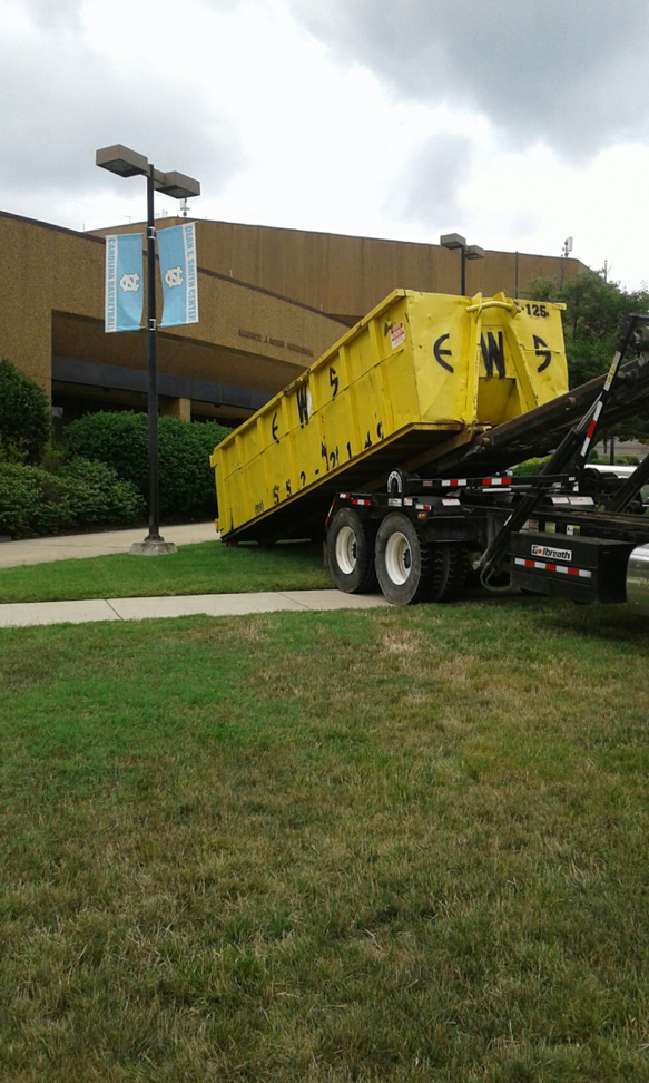 An Elite Waste Services roll-off truck delivering a yellow dumpster for junk removal in Raleigh, NC.