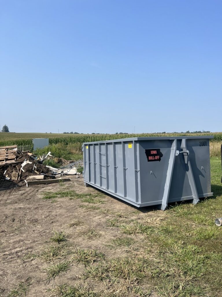 A grey roll-off dumpster from Iowa Roll Off placed next to a large pile of debris and junk in a field in Ankeny, IA.