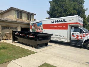 An empty roll-off dumpster placed next to a U-Haul truck in a residential driveway by Bakersfield Roll-Off Service in Bakersfield, CA.