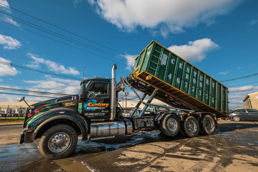 A Century Waste Management truck loading a large green roll-off dumpster for junk removal in Sterling Heights, MI.