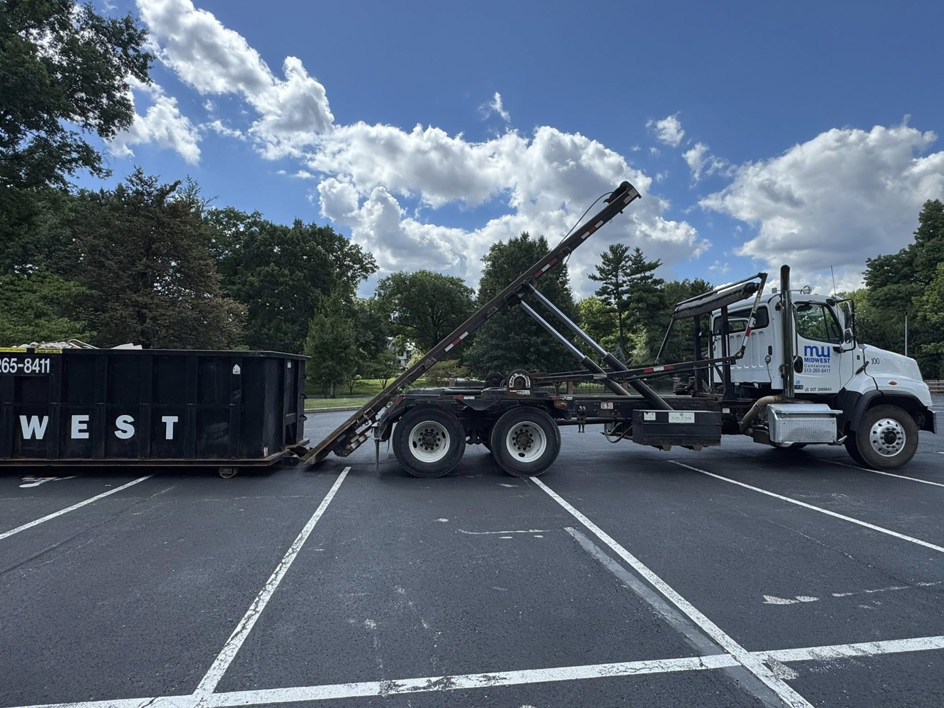 A Midwest Containers roll-off dumpster truck loading a black dumpster in a parking lot in Cincinnati, OH.