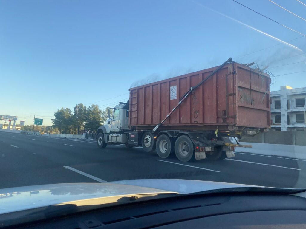 A Waste Connections roll-off dumpster truck transporting a large container full of debris on a highway in Houston, TX.