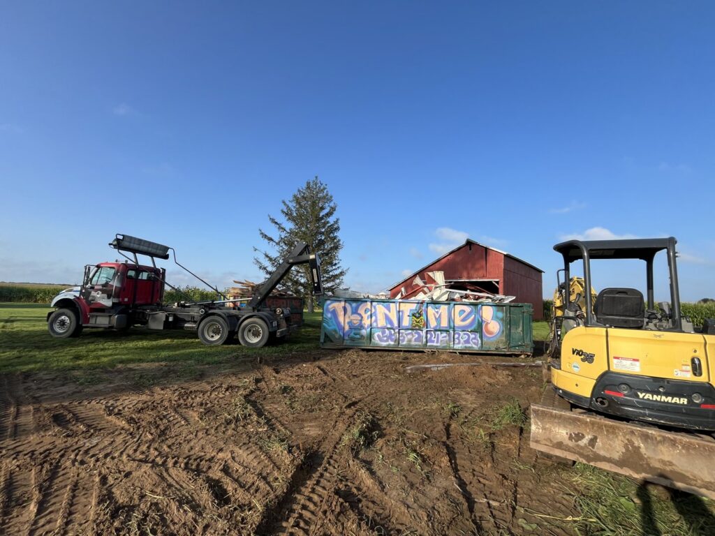 A roll-off truck with a green debris dumpster and an excavator from AT Disposal at a rural site in Dewitt, IA.