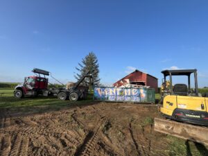 A roll-off truck with a green debris dumpster and an excavator from AT Disposal at a rural site in Dewitt, IA.