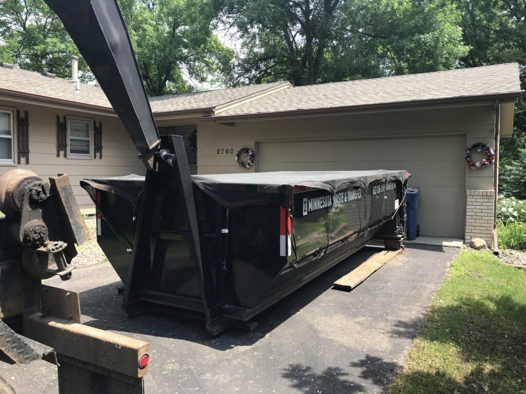 A roll-off truck from Minnesota Waste and Transfer deploying a covered dumpster in a residential driveway in Champlin, MN