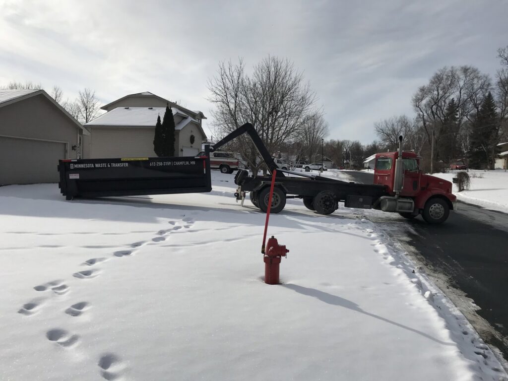 A red roll-off truck from Minnesota Waste and Transfer with a black dumpster on a snowy residential street in Champlin, MN