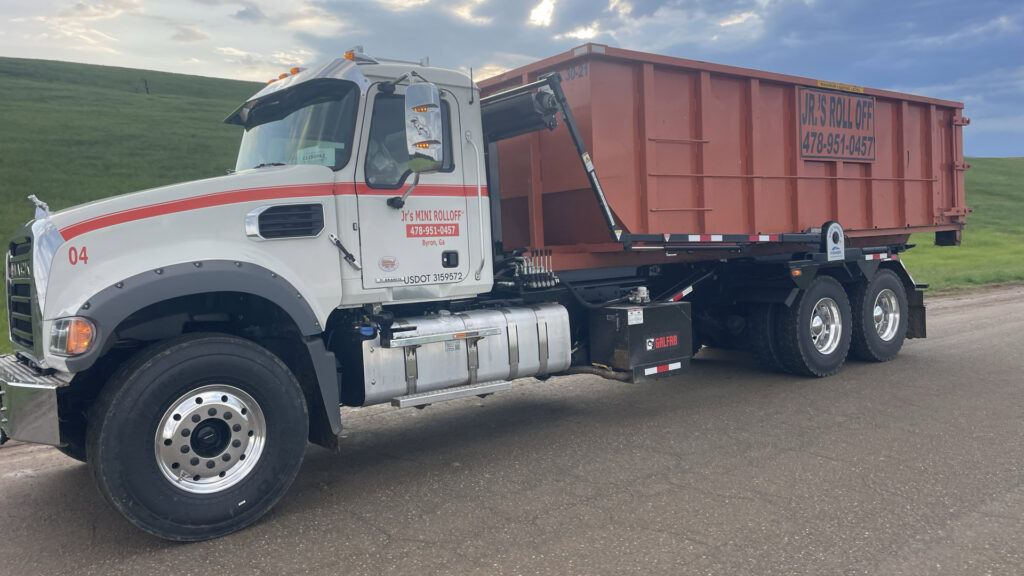 A Jr's Mini Roll Off LLC truck with an orange roll-off dumpster parked on a road in Byron, GA.