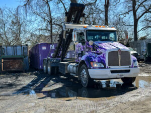 A JP's Disposal Inc. roll-off truck with a purple dumpster in a muddy lot in Hampton, VA.