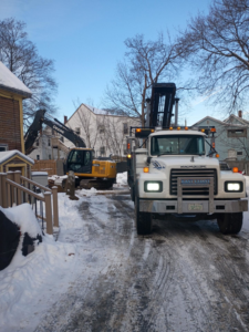 A roll-off truck and excavator on a snowy residential job site for East Coast Container Services LLC in Northwood, NH.