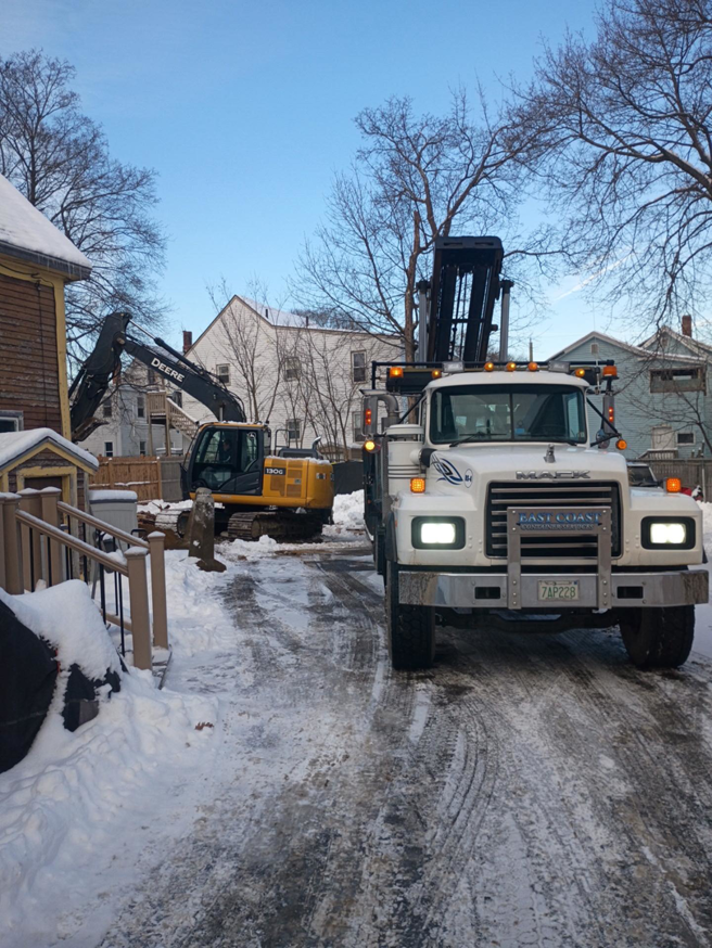 A roll-off truck and excavator on a snowy residential job site for East Coast Container Services LLC in Northwood, NH.