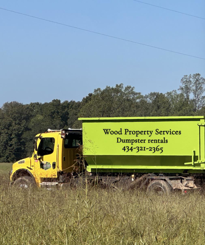 A yellow roll-off truck with a green dumpster from Wood Property Services & Dumpster Rental LLC in a field in Wilmington, NC.