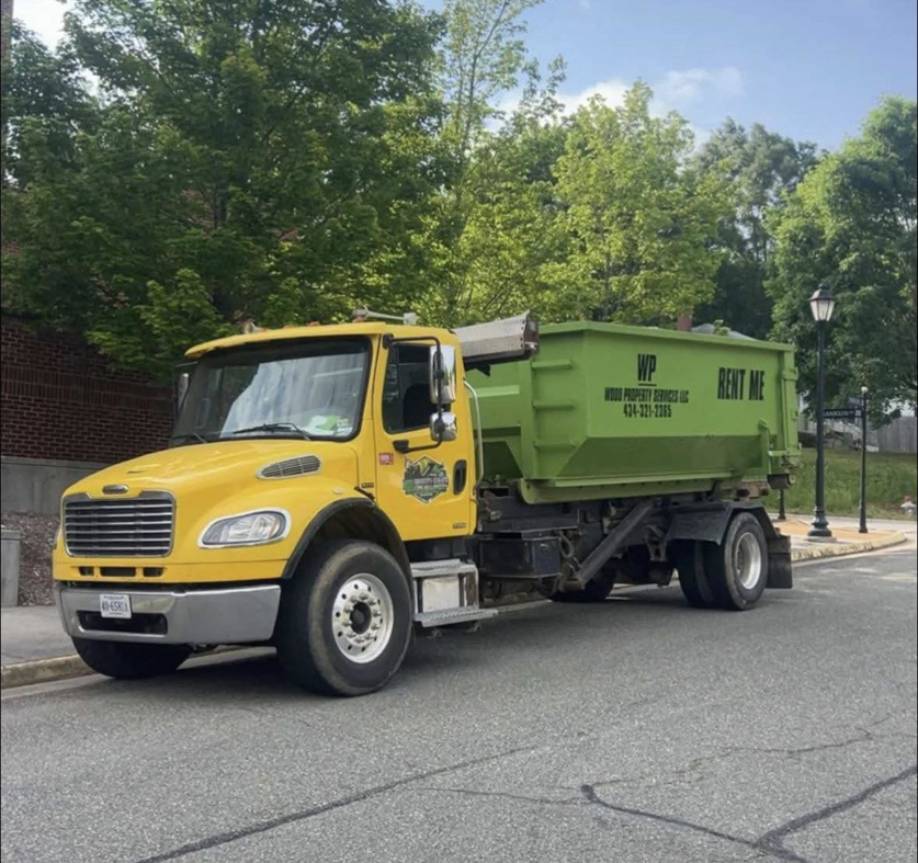 A yellow roll-off truck with a green dumpster from Wood Property Services & Dumpster Rental LLC parked on a street in Wilmington, NC.