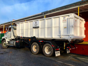 A roll-off truck with a large white dumpster parked next to a commercial building, ready for waste removal by Standard Waste Enterprises in Greenville, SC.