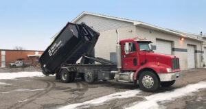 A red roll-off truck from Minnesota Waste and Transfer lifting a black dumpster in front of a commercial building in Champlin, MN