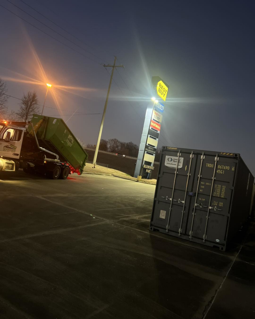 A Top Line Dumpsters LLC roll-off truck lifting a green dumpster at a Best Buy location at night in Hammond, LA.