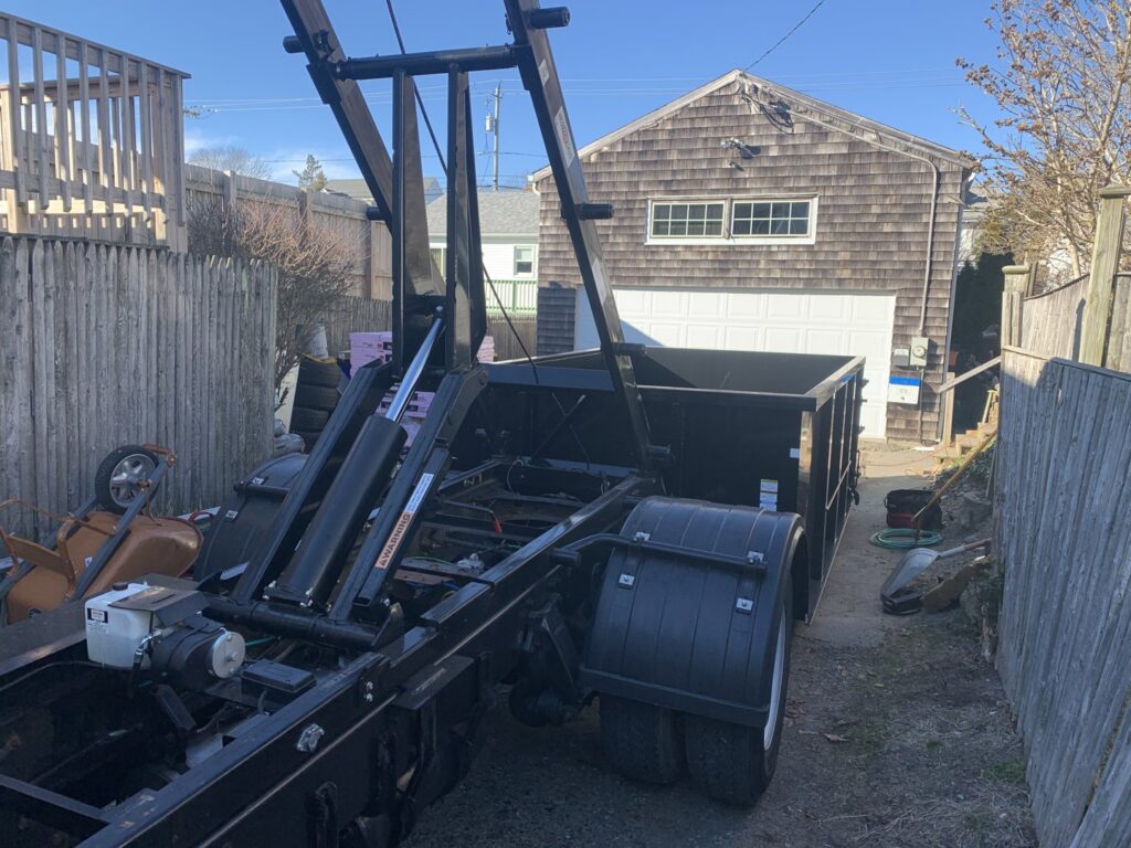 A roll-off truck placing a black dumpster in a residential backyard for junk removal services from Convenient Disposal & Sanitation in Fall River, MA.