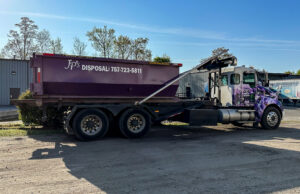A JP's Disposal Inc. roll-off truck with a purple dumpster in an industrial area in Hampton, VA.