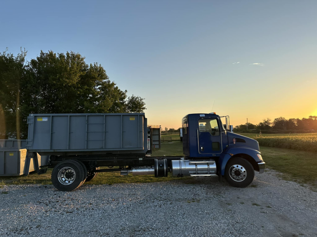 A blue roll-off truck with an empty dumpster on its bed, parked at sunset for Iowa Roll Off in Ankeny, IA.