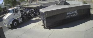 A Payless Rolloff truck delivering or picking up a covered roll-off dumpster on a paved area in Albuquerque, NM.
