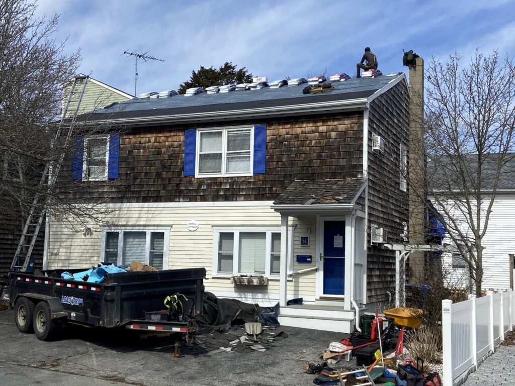 A roofing crew performing shingle replacement on a house, with a dumpster for debris, by Eagle Exteriors in Cumberland, RI.