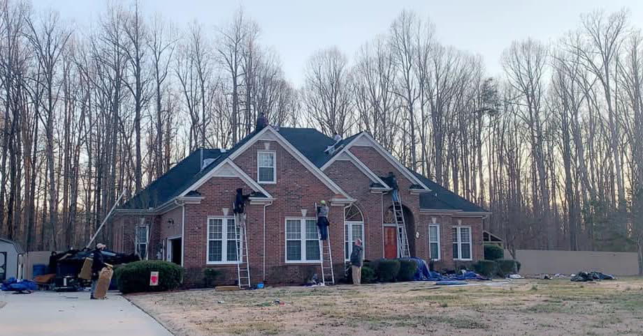 Workers on ladders performing a roofing job on a brick house for First Class Roofing Inc. in High Point, NC