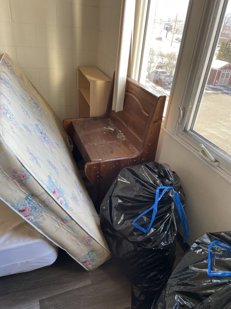 A room corner with old mattresses, a wooden bench, and black trash bags ready for junk removal by Missoula Junk Removal, LLC in Missoula, MT.