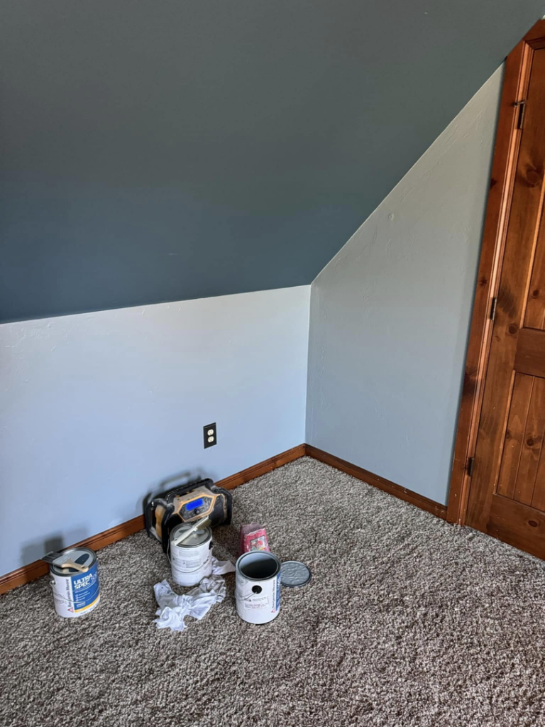 A room painting project with an angled ceiling and walls, showing paint cans on the carpet, by Sanders Construction & Handyman Services, LLC in Oklahoma City, OK.