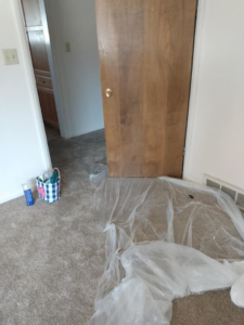 A room prepared for handyman services with plastic sheeting on the carpet and a wooden door propped up by Bevard Enterprises in Millcreek, UT.