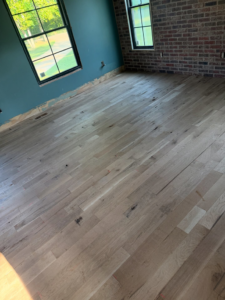 A room with an unfinished hardwood floor, large windows, and a brick accent wall by Cordell Hardwood Flooring in Lexington, KY.