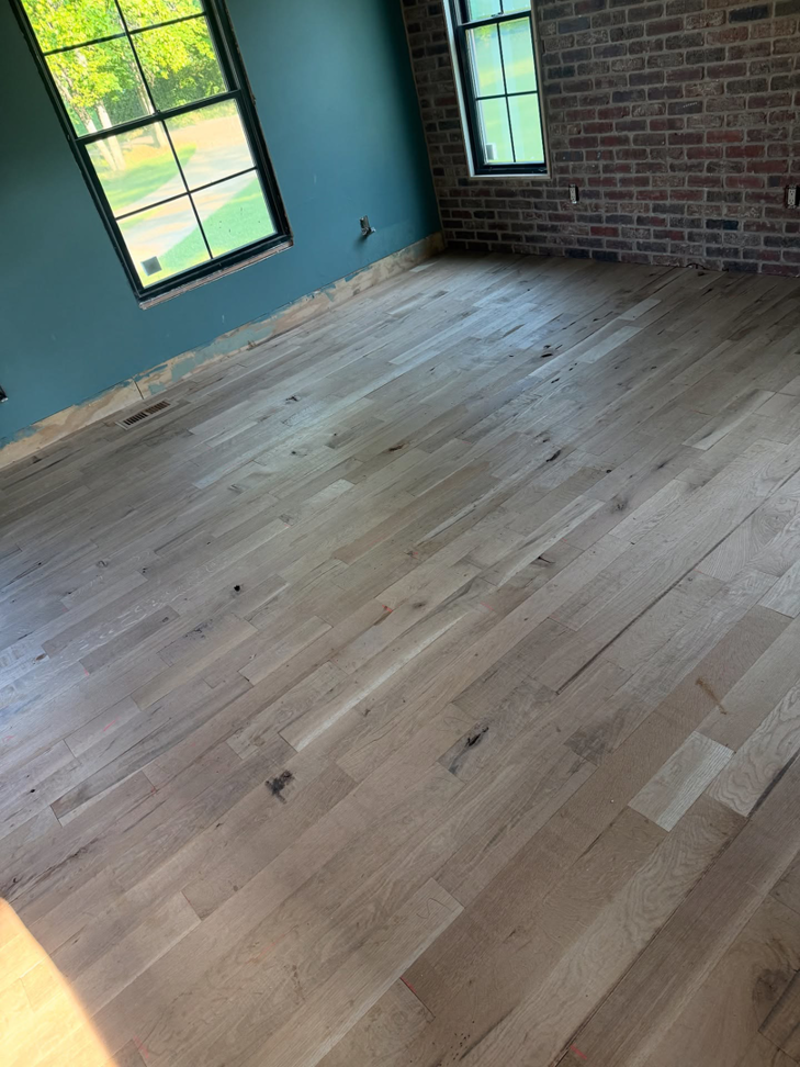 A room with an unfinished hardwood floor, large windows, and a brick accent wall by Cordell Hardwood Flooring in Lexington, KY.