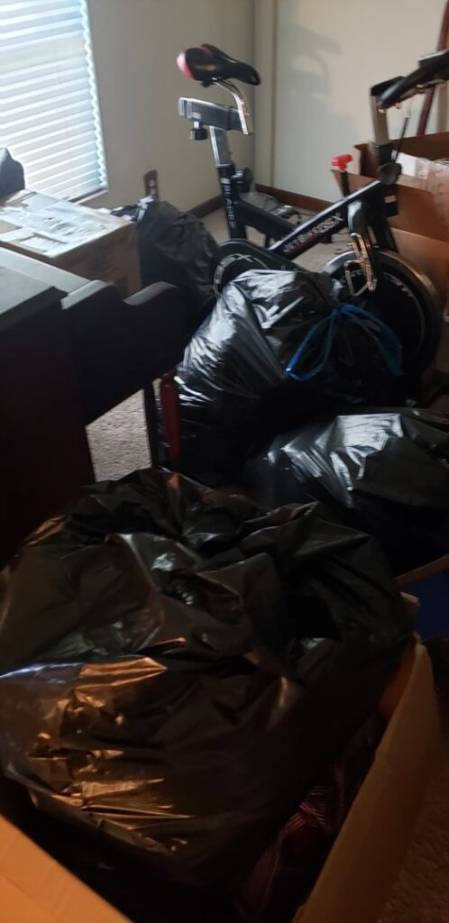 A room filled with black trash bags, boxes, and an exercise bike, indicating a cleanout service by Junk Fade Away, LLC in Jacksonville, FL.