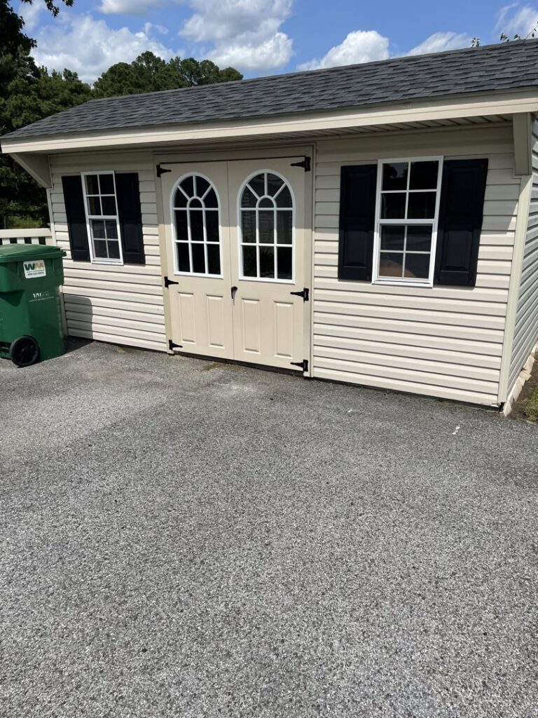 A newly constructed or renovated shed with light-colored siding, double doors, and black shutters by Shore Home Construction Roofing in Salisbury, MD.