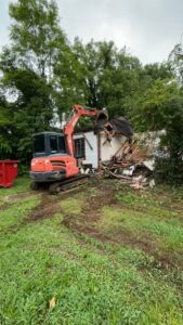 An excavator demolishing a shed with a dumpster for junk removal by Green Brothers Services in Harvest, AL