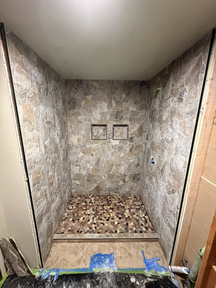 A shower stall under construction with stone-look wall panels and a pebble floor by Connolly Tile LLC in Medford, OR.