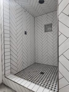 A newly tiled shower featuring white herringbone wall tiles and black penny floor tiles by D E Custom Floors in Waukesha, WI.